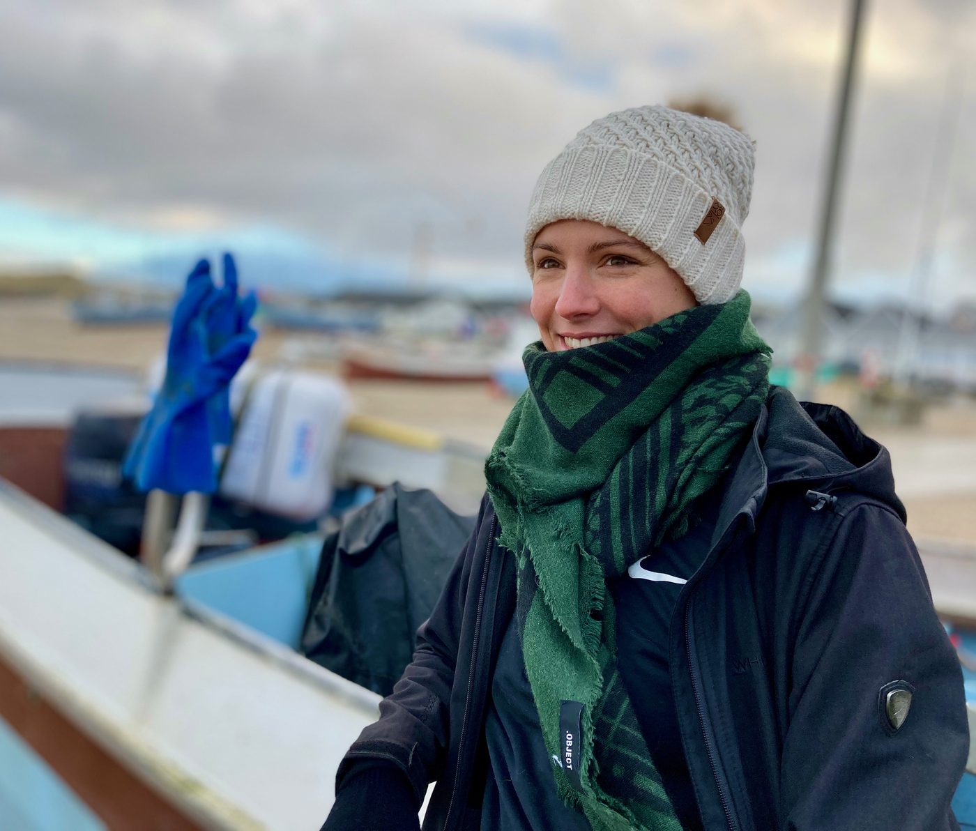 Sillan smiler på havnen en blæsende dag, med hue og tørklæde og joller i baggrunden.